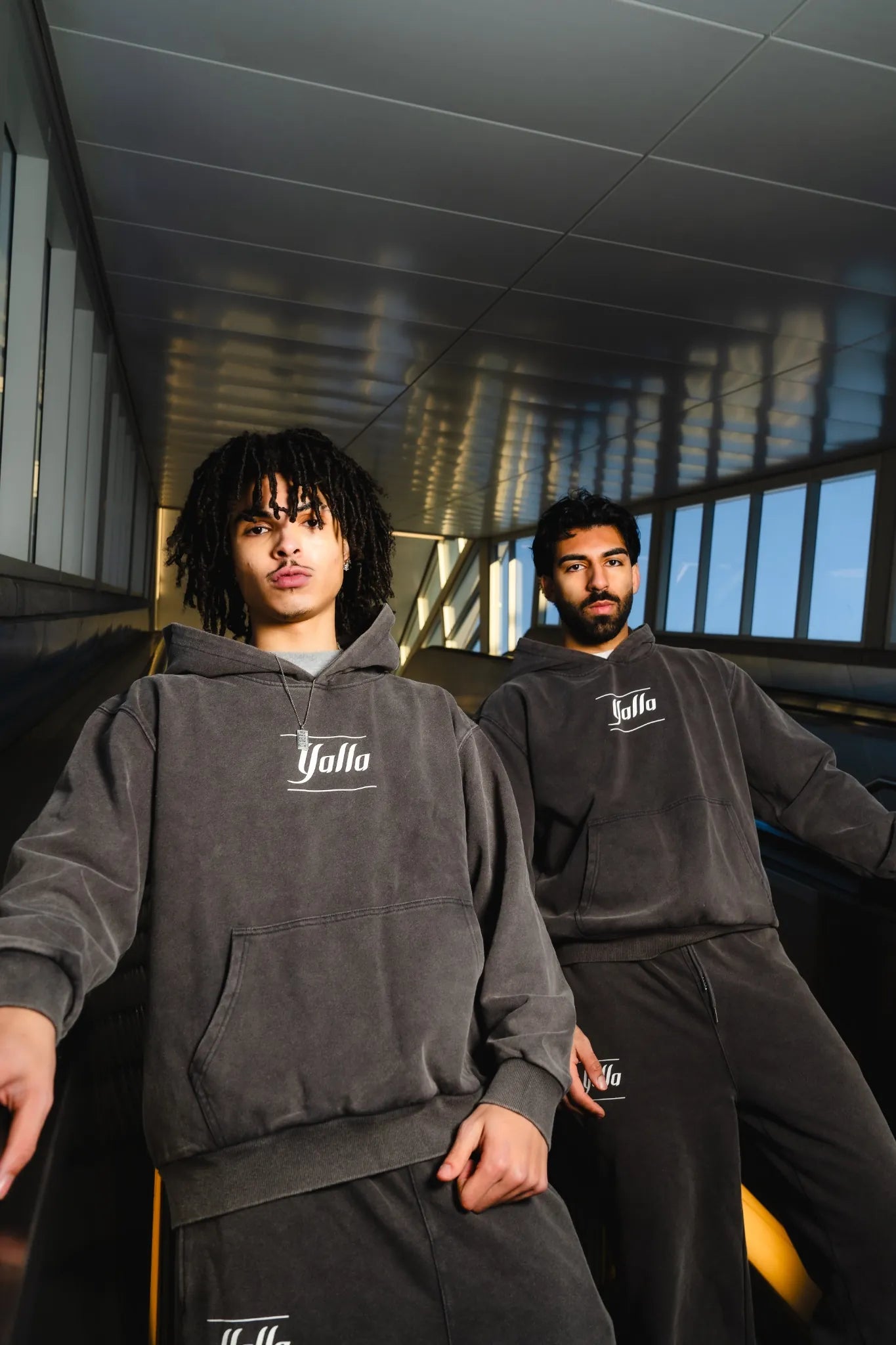 Two male models wearing heavyweight dark grey Arab tracksuits by Yalla Apparel, featuring soft cotton fabric and minimalist embroidered logos.