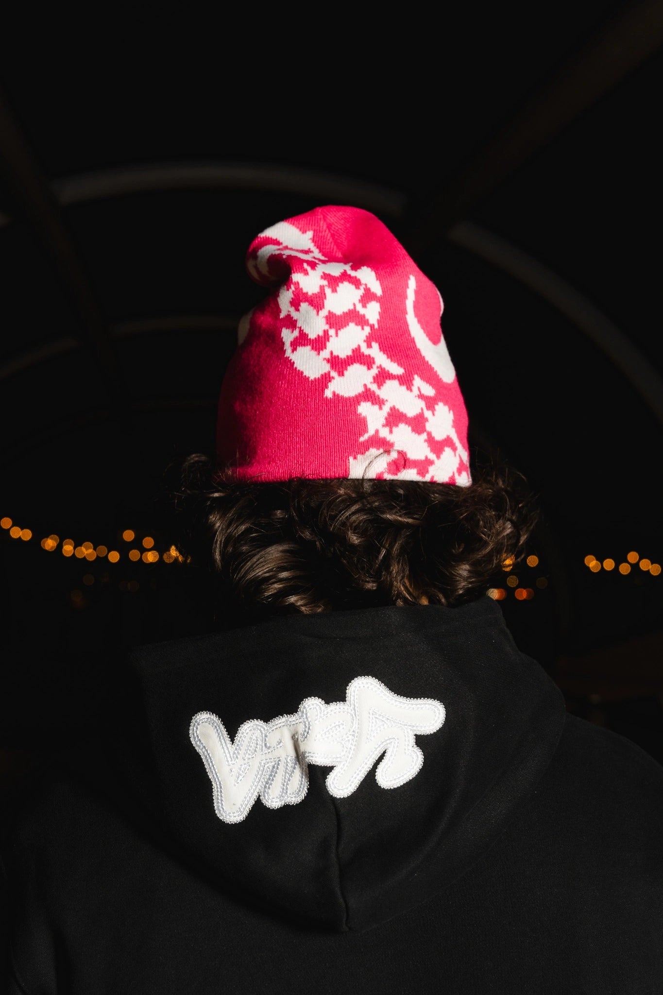 Back view of a model wearing a pink Kufiya beanie and a black Yalla hoodie at night. Arab-inspired streetwear featuring Kufiya patterns and Arabic calligraphy. A fusion of modern fashion and cultural heritage.