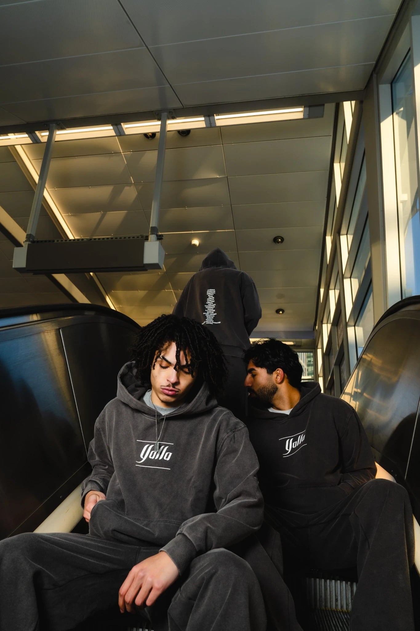 Two people sitting on an escalator wearing gray hoodies with a brand logo showing the Arab Tracksuit. 